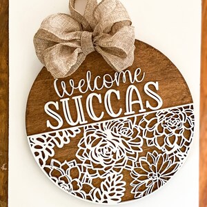 Peut inclure: Une pancarte ronde en bois avec un n&oelig;ud en toile de jute sur le dessus. La pancarte indique "Welcome Succas" en lettres blanches. La moiti&eacute; inf&eacute;rieure de la pancarte est d&eacute;cor&eacute;e d'un motif d&eacute;coup&eacute; blanc de succulentes.