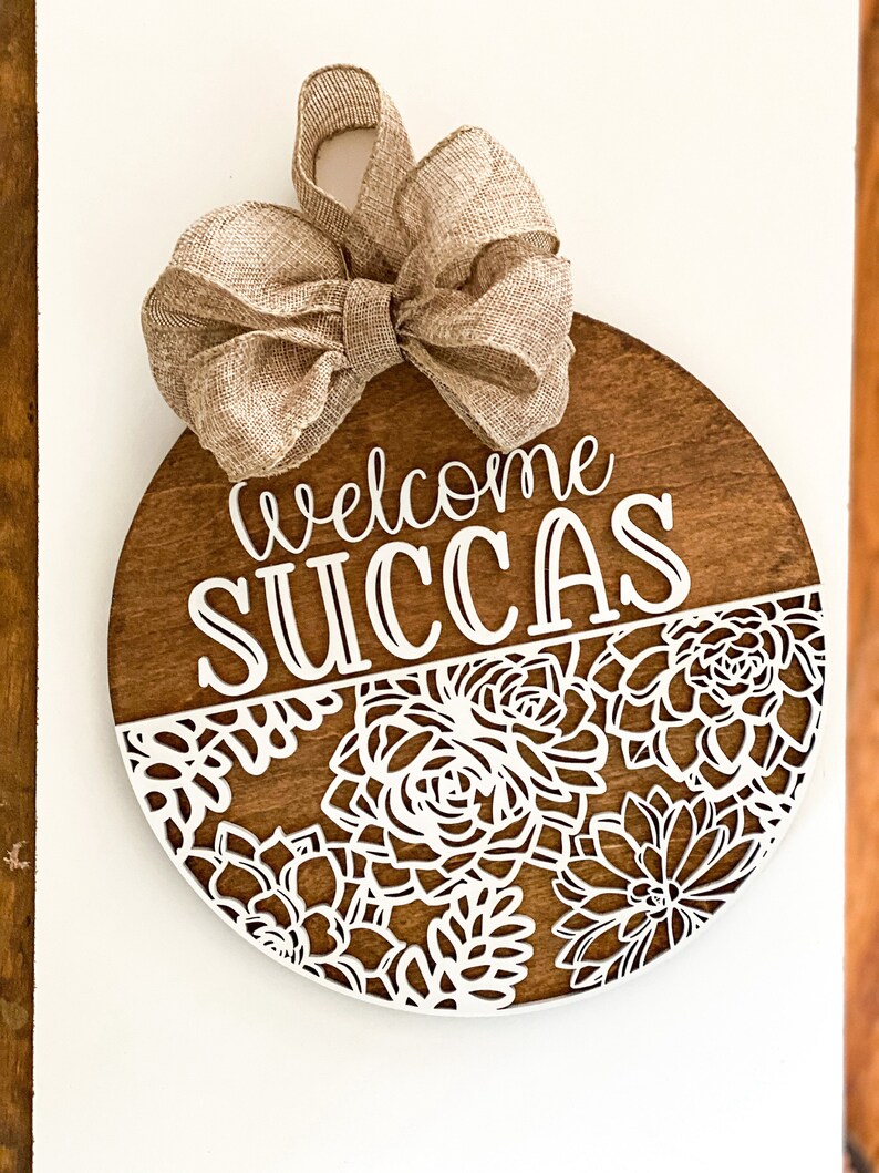 Peut inclure: Une pancarte ronde en bois avec un n&oelig;ud en toile de jute sur le dessus. La pancarte indique "Welcome Succas" en lettres blanches. La moiti&eacute; inf&eacute;rieure de la pancarte est d&eacute;cor&eacute;e d'un motif d&eacute;coup&eacute; blanc de succulentes.