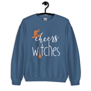 Op de afbeelding: Een blauwe sweater met ronde hals en de witte tekst cheers witches. Het ontwerp bevat een oranje heksenhoed boven het woord cheers en een oranje bezemsteel die de letter i in het woord witches vervangt op het midden van de voorzijde.