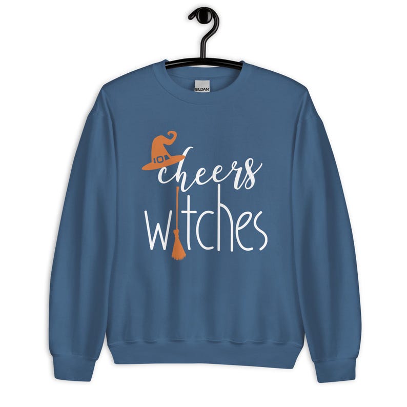 Op de afbeelding: Een blauwe sweater met ronde hals en de witte tekst cheers witches. Het ontwerp bevat een oranje heksenhoed boven het woord cheers en een oranje bezemsteel die de letter i in het woord witches vervangt op het midden van de voorzijde.