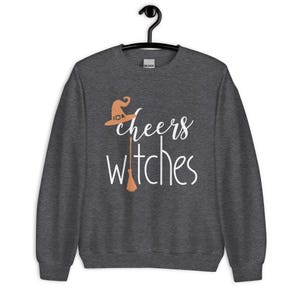 Op de afbeelding: Een donkergrijs gem&ecirc;leerd sweatshirt met lange mouwen en de witte tekst "cheers witches" op de voorzijde. Het ontwerp bevat een lichtoranje heksenhoed boven het woord cheers en een lichtoranje bezemsteel die de letter t vervangt.
