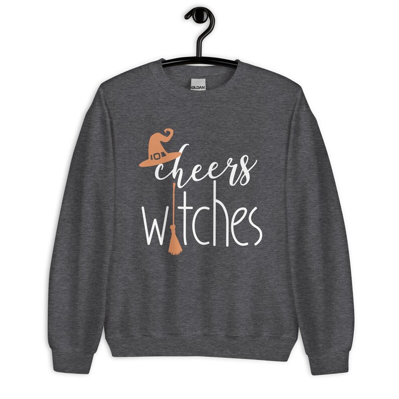 Op de afbeelding: Een donkergrijs gem&ecirc;leerd sweatshirt met lange mouwen en de witte tekst "cheers witches" op de voorzijde. Het ontwerp bevat een lichtoranje heksenhoed boven het woord cheers en een lichtoranje bezemsteel die de letter t vervangt.
