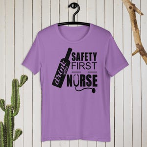 K&ouml;nnte beinhalten: Lilafarbenes T-Shirt mit einer schwarzen Grafik einer Weinflasche und dem Text "drink with a nurse" und "safety first".