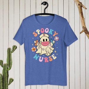 以下が含まれることがあります： 「Spooky Nurse」のグラフィックがプリントされた青いTシャツ。看護師の帽子とマスクをつけたお化けが、包帯と注射器を持っています。テキストはカラフルなレトロスタイルの文字で、花と星が描かれています。