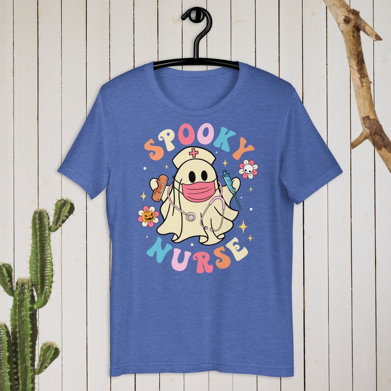 以下が含まれることがあります： 「Spooky Nurse」のグラフィックがプリントされた青いTシャツ。看護師の帽子とマスクをつけたお化けが、包帯と注射器を持っています。テキストはカラフルなレトロスタイルの文字で、花と星が描かれています。