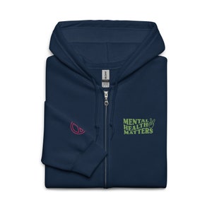 May include: A navy blue zip-up hoodie with a green and yellow embroidered design that says "Mental Health Matters" and a peace sign. The hoodie also has a pink logo on the left side of the chest.