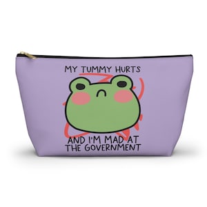 May include: A lavender cosmetic bag with a gold zipper. The bag features a green frog face with pink cheeks and the text "MY TUMMY HURTS AND I'M MAD AT THE GOVERNMENT" in black and red.