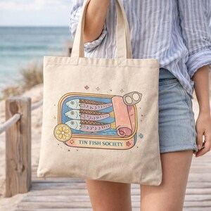 Woman carrying sardine tote bag with Tin Fish Society design walking on beach boardwalk