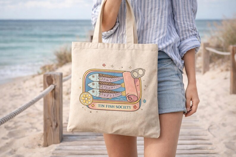 Woman carrying sardine tote bag with Tin Fish Society design walking on beach boardwalk