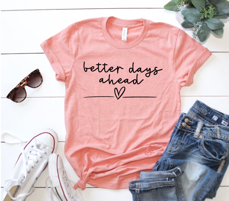 May include: A peach-coloured t-shirt with the text "better days ahead" in black script with a small heart underneath. The shirt is laid out on a white wooden surface with a pair of sunglasses and white trainers.