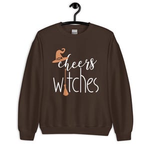 Op de afbeelding: Een donkerbruin sweatshirt met lange mouwen en de witte tekst "cheers witches" op de voorzijde. Het ontwerp bevat een lichtoranje heksenhoed boven het woord cheers en een lichtoranje bezemsteel die de letter t in witches vervangt.