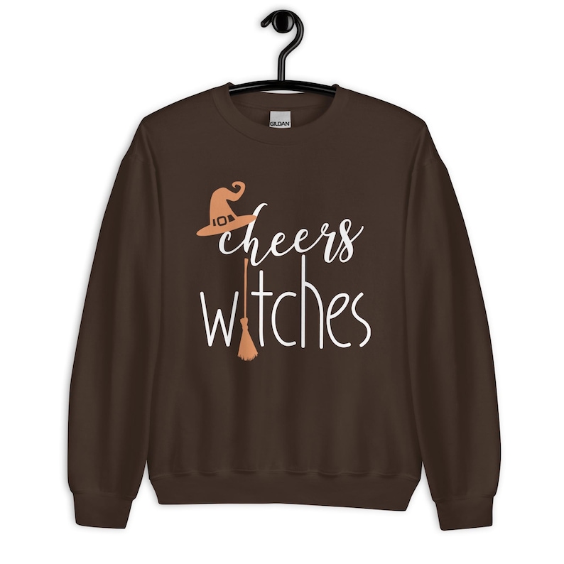 Op de afbeelding: Een donkerbruin sweatshirt met lange mouwen en de witte tekst "cheers witches" op de voorzijde. Het ontwerp bevat een lichtoranje heksenhoed boven het woord cheers en een lichtoranje bezemsteel die de letter t in witches vervangt.