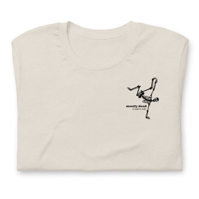 Puede incluir: Una camiseta beige con un gr&aacute;fico de esqueleto negro y el texto "mostly dead is slightly alive".