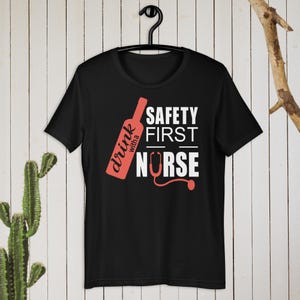K&ouml;nnte beinhalten: Schwarzes T-Shirt mit einer roten Weinflasche und dem Text "drink with a nurse" und "safety first" in Wei&szlig;.