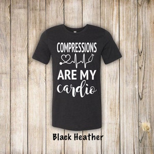 May include: Black heather grey t-shirt with white text that reads "COMPRESSIONS ARE MY cardio" with a heart and EKG line graphic.