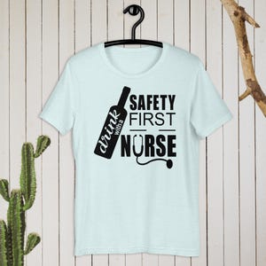 K&ouml;nnte beinhalten: Hellblaues T-Shirt mit einer schwarzen Grafik einer Weinflasche und dem Text "drink with a nurse" und "safety first" mit einem Stethoskop.