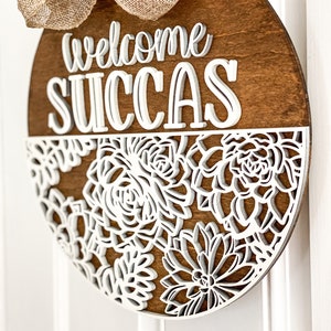 Peut inclure: Une suspension de porte ronde en bois avec les mots "welcome succas" en lettres blanches en relief. La moiti&eacute; inf&eacute;rieure de la suspension est d&eacute;cor&eacute;e d'un motif d&eacute;coup&eacute; blanc de succulentes.
