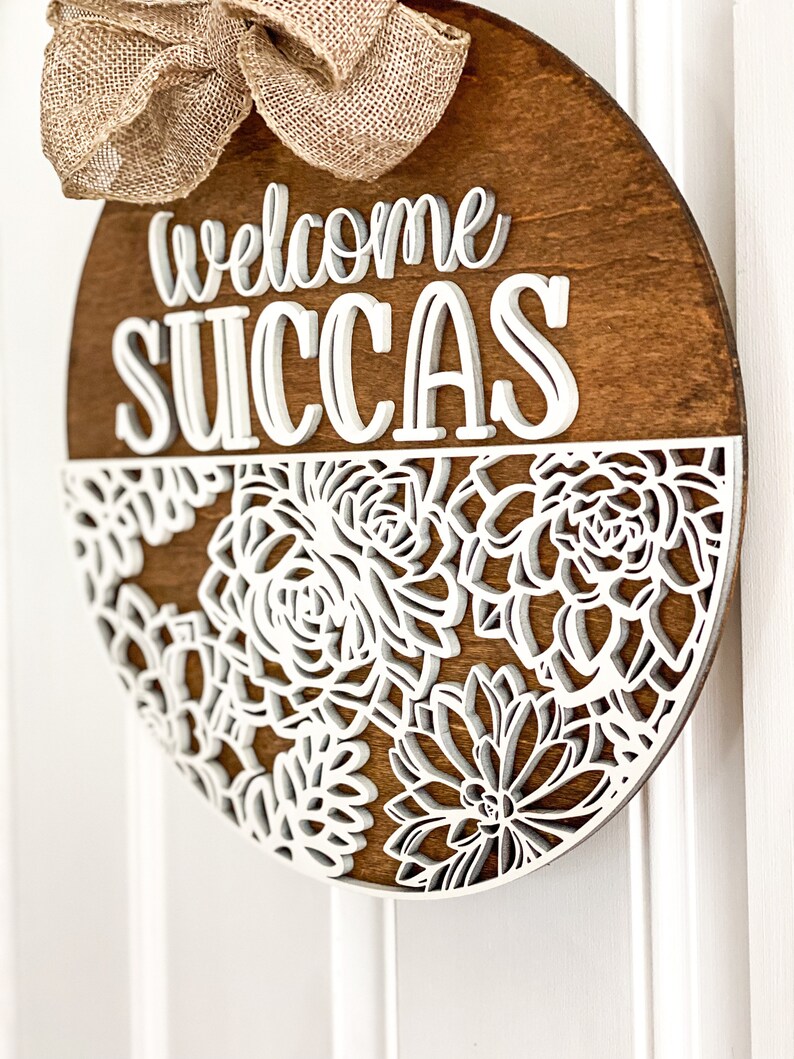 Peut inclure: Une suspension de porte ronde en bois avec les mots "welcome succas" en lettres blanches en relief. La moiti&eacute; inf&eacute;rieure de la suspension est d&eacute;cor&eacute;e d'un motif d&eacute;coup&eacute; blanc de succulentes.