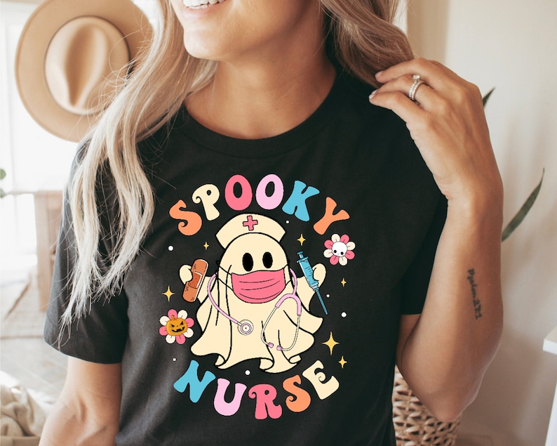 以下が含まれることがあります： 「Spooky Nurse」と書かれたカラフルなグラフィックが特徴の黒いTシャツ。看護師の帽子とマスクを着用し、注射器と包帯を持っている幽霊のデザインです。デザインには花と星が含まれています。