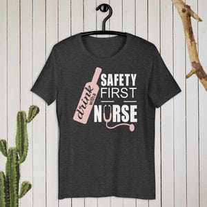 K&ouml;nnte beinhalten: Dunkelgraues T-Shirt mit einem pinkfarbenen Wein-Flaschen-Motiv und dem Text "drink with a nurse" und "safety first" in Wei&szlig;.