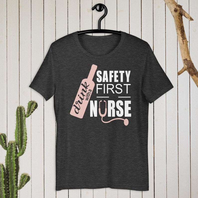 K&ouml;nnte beinhalten: Dunkelgraues T-Shirt mit einem pinkfarbenen Wein-Flaschen-Motiv und dem Text "drink with a nurse" und "safety first" in Wei&szlig;.