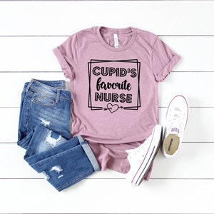 May include: A mauve t-shirt with the text "CUPID'S favorite NURSE" in a black square frame with a heart and arrow.