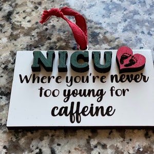K&ouml;nnte beinhalten: Wei&szlig;er Holzanh&auml;nger mit dem Text "NICU Where you're never too young for caffeine" in schwarzer Schrift. Ein rotes Herz mit einem Fu&szlig;abdruckmuster befindet sich auf der rechten Seite des Anh&auml;ngers.