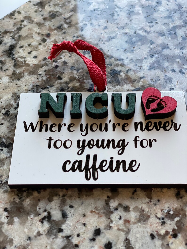 K&ouml;nnte beinhalten: Wei&szlig;er Holzanh&auml;nger mit dem Text "NICU Where you're never too young for caffeine" in schwarzer Schrift. Ein rotes Herz mit einem Fu&szlig;abdruckmuster befindet sich auf der rechten Seite des Anh&auml;ngers.