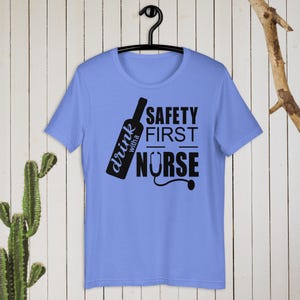 K&ouml;nnte beinhalten: Ein hellblaues T-Shirt mit einem schwarzen Grafikdesign. Die Grafik zeigt eine Weinflasche mit dem Text "drink with a nurse" und den Worten "safety first" dar&uuml;ber. Ein Stethoskop ist im Design enthalten.