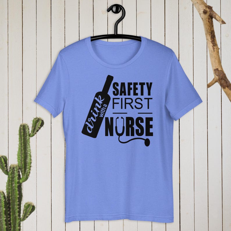 K&ouml;nnte beinhalten: Ein hellblaues T-Shirt mit einem schwarzen Grafikdesign. Die Grafik zeigt eine Weinflasche mit dem Text "drink with a nurse" und den Worten "safety first" dar&uuml;ber. Ein Stethoskop ist im Design enthalten.
