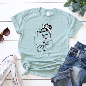 Op de afbeelding: Een lichtblauw t-shirt met een afbeelding van een vrouw met een stethoscoop en de tekst "ER Nurse".