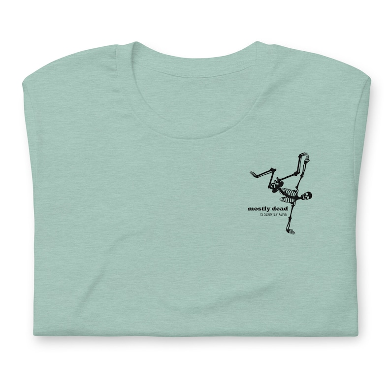 Puede incluir: Una camiseta azul claro con un gr&aacute;fico de esqueleto negro haciendo un movimiento de breakdance. El texto "mostly dead is slightly alive" est&aacute; impreso debajo del esqueleto.