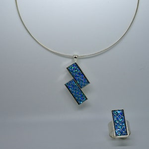 Peut inclure: Un collier en argent avec un pendentif bleu iridescent et une bague assortie. Le pendentif est composé de deux pièces rectangulaires reliées à un angle. La bague a une pierre rectangulaire bleu iridescent.