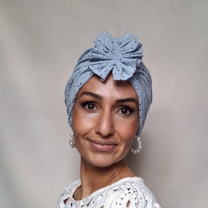 Könnte beinhalten: Hellblaues Turban mit floralem Akzent. Der Turban besteht aus einem perforierten Stoff. Die Person trägt ein weißes Oberteil mit offenem Webmuster und Perlenohrringe.