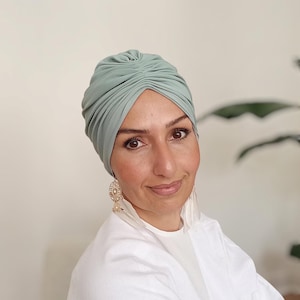 Könnte beinhalten: Ein salbeigrüner Turban mit gerafftem Design. Die Kopfbedeckung besteht aus einem weichen Stoff. Die Person trägt ein weißes Oberteil und goldene Ohrringe.