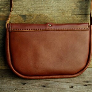 Może przedstawiać: Brązowa sk&oacute;rzana torba na ramię z klapką i jednym paskiem. Torba ma zaokrąglony kształt i gładkie wykończenie ze sk&oacute;ry.