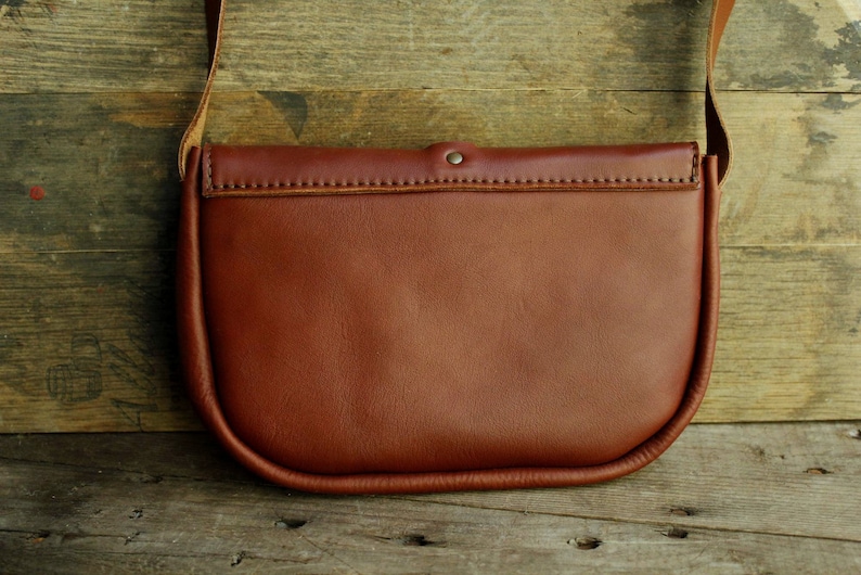 Może przedstawiać: Brązowa sk&oacute;rzana torba na ramię z klapką i jednym paskiem. Torba ma zaokrąglony kształt i gładkie wykończenie ze sk&oacute;ry.