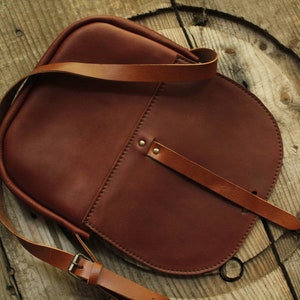 Może przedstawiać: Brązowa sk&oacute;rzana torba na ramię z długim, regulowanym paskiem. Torba ma klapę z klamrą i małą kieszeń z przodu.