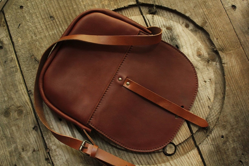 Może przedstawiać: Brązowa sk&oacute;rzana torba na ramię z długim, regulowanym paskiem. Torba ma klapę z klamrą i małą kieszeń z przodu.