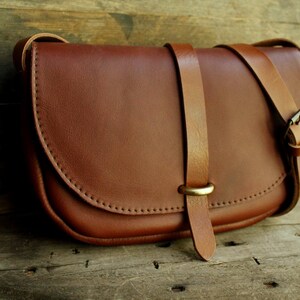 Może przedstawiać: Brązowa sk&oacute;rzana torebka na ramię z klapką i klamrą mosiężną. Torebka ma długi, regulowany pasek.