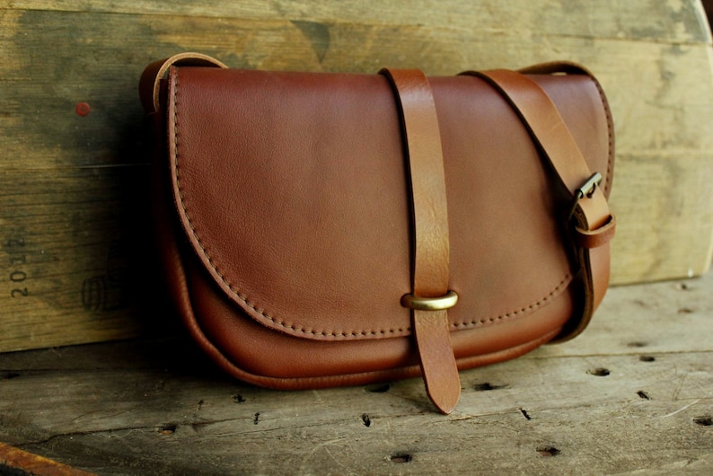 Może przedstawiać: Brązowa sk&oacute;rzana torebka na ramię z klapką i klamrą mosiężną. Torebka ma długi, regulowany pasek.