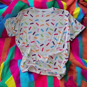 May include: White t-shirt with a colorful sprinkle pattern. The shirt is laid out on a rainbow striped blanket.