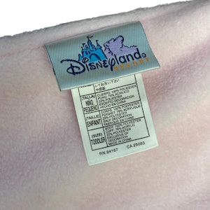May include: A light pink blanket with a Disneyland Resort tag. The tag displays the Disneyland logo and text in multiple languages. The blanket is made of polyester and acrylic. The tag also includes washing instructions.