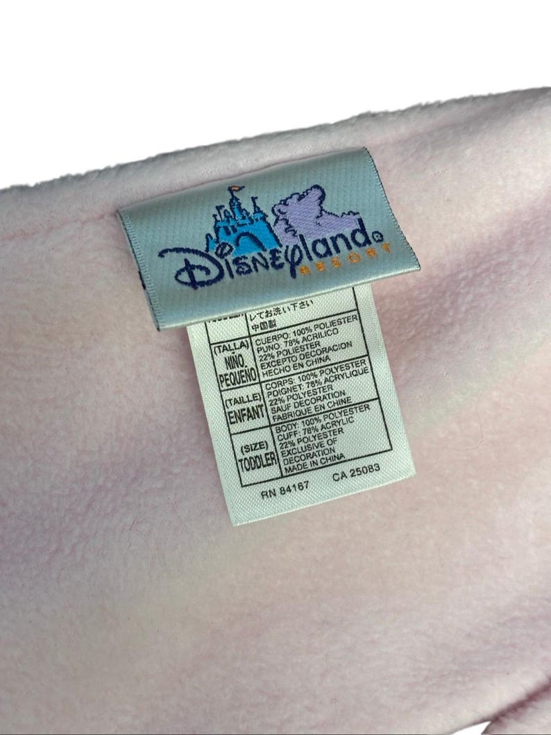 May include: A light pink blanket with a Disneyland Resort tag. The tag displays the Disneyland logo and text in multiple languages. The blanket is made of polyester and acrylic. The tag also includes washing instructions.