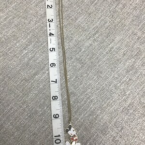 Puede incluir: Un collar de cadena dorada con un dije de Mickey Mouse. El dije es blanco con contornos negros y pantalones cortos rojos. La cadena mide aproximadamente 25 cm de largo.