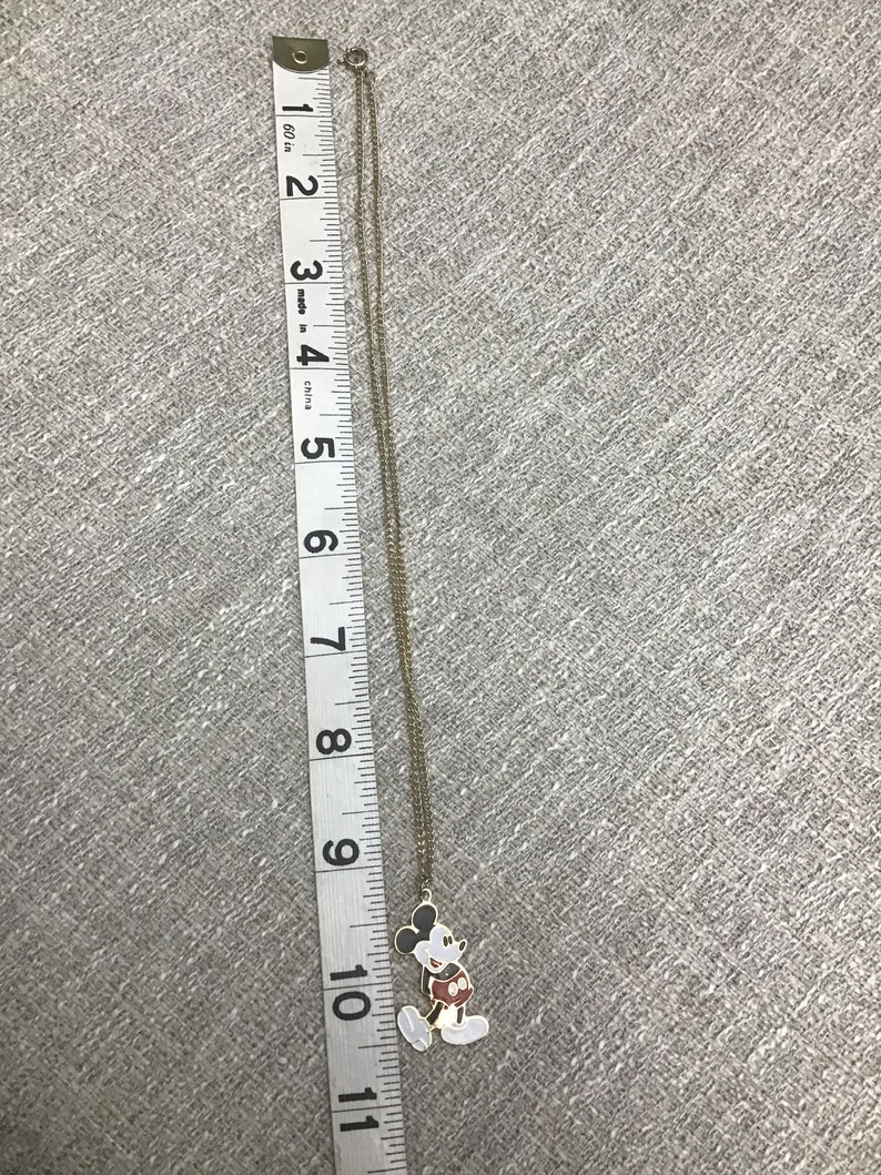 Puede incluir: Un collar de cadena dorada con un dije de Mickey Mouse. El dije es blanco con contornos negros y pantalones cortos rojos. La cadena mide aproximadamente 25 cm de largo.