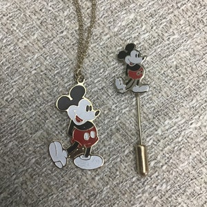 Puede incluir: Un collar de cadena de oro y un pin con Mickey Mouse. Ambos son de color dorado y presentan una imagen en blanco y negro de Mickey Mouse con pantalones cortos rojos.