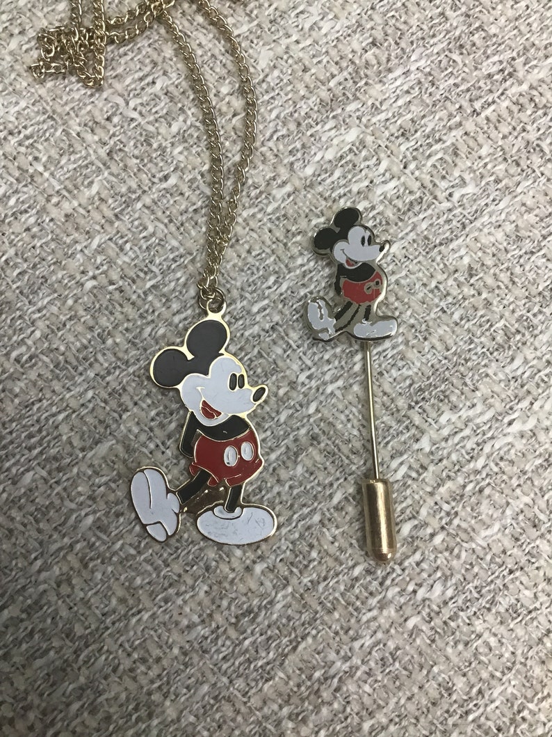 Puede incluir: Un collar de cadena de oro y un pin con Mickey Mouse. Ambos son de color dorado y presentan una imagen en blanco y negro de Mickey Mouse con pantalones cortos rojos.