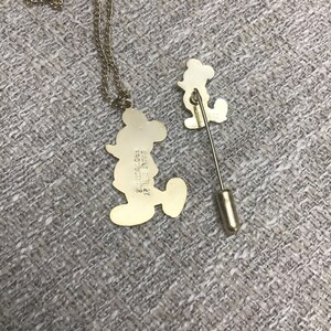 Puede incluir: Un conjunto de collar y broche de Mickey Mouse en tono dorado. El collar presenta un colgante de Mickey Mouse con el texto "Walt Disney Productions" en la parte posterior. El broche es un dise&ntilde;o de Mickey Mouse m&aacute;s peque&ntilde;o con un cierre de broche en tono dorado.