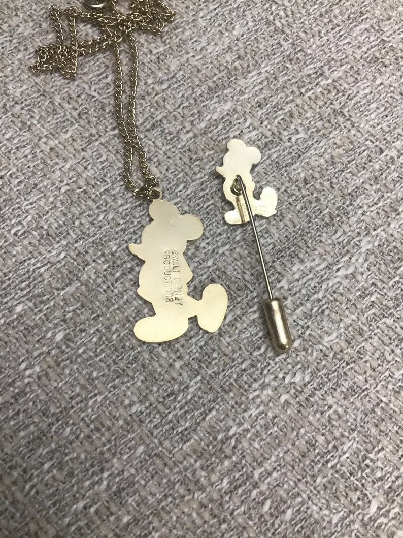 Puede incluir: Un conjunto de collar y broche de Mickey Mouse en tono dorado. El collar presenta un colgante de Mickey Mouse con el texto "Walt Disney Productions" en la parte posterior. El broche es un dise&ntilde;o de Mickey Mouse m&aacute;s peque&ntilde;o con un cierre de broche en tono dorado.